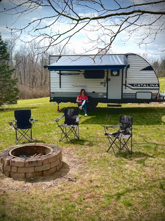 A woman relaxing by a Coachmen Catalina RV rental, with a fire pit and chairs in the grassy outdoor space.. Forest River Coachmen Catalina Summit 2024