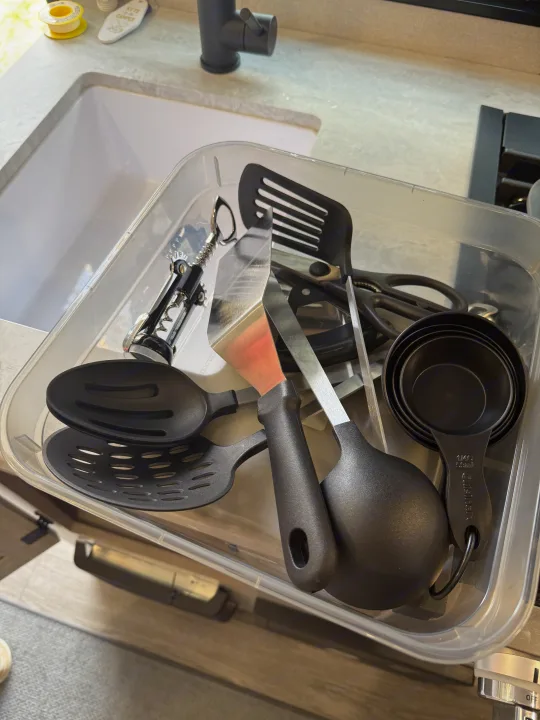 RV rental kitchen: a clear bin filled with essential black cooking utensils including spatulas, spoons, measuring cups, and a corkscrew.. Forest River Coachmen Catalina Summit 2024