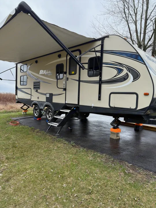 A tan Keystone Bullet RV rental is set up at a damp campsite with its awning extended.. Keystone RV Bullet Ultra Lite 2017