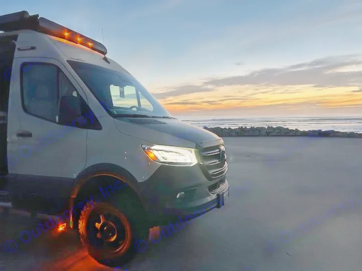 RV rental camper van parked on a dark beach at sunset. Its lights glow, facing the ocean.. Winnebago Revel 2024