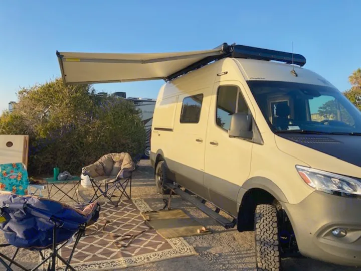 Perfect day at a campsite! This rv rental, a cream-colored camper van with its awning out, offers comfortable outdoor living.. Winnebago Revel 2024