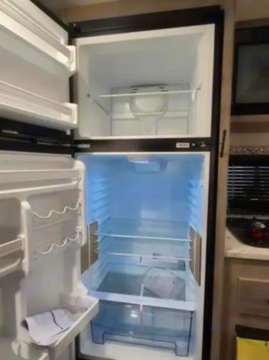 Inside an RV rental: a clean, empty refrigerator/freezer unit, with a partial kitchen visible to the right.. Dutchmen Coleman Lantern LT 2021