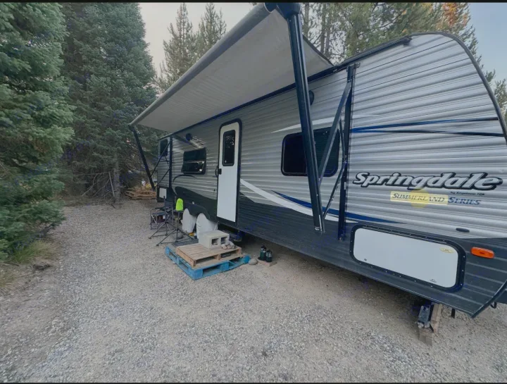 Spacious Springdale Summerland RV rental setup on a gravel campsite with extended awning amidst tall trees. Ideal for outdoor living.. Keystone RV Summerland 2017