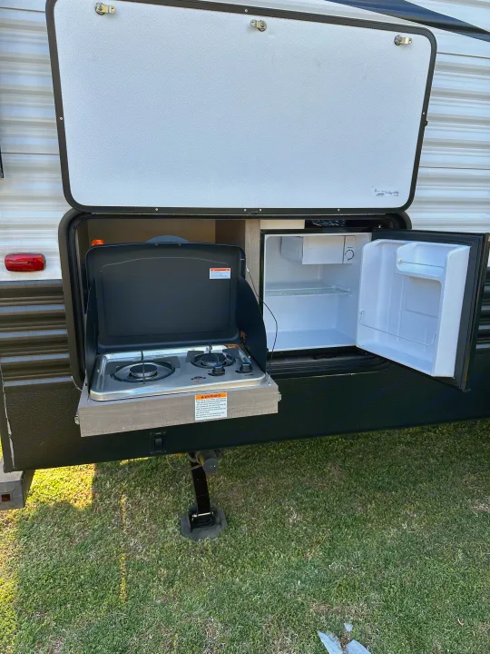 RV rental outdoor kitchen setup with a two-burner stove and mini-fridge open, showing shelves. On grass.. Keystone Hideout 2021
