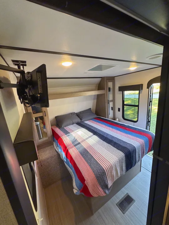 Inside a comfy RV rental bedroom: striped bed, mounted TV, and window with a scenic view.. Keystone Hideout 2021