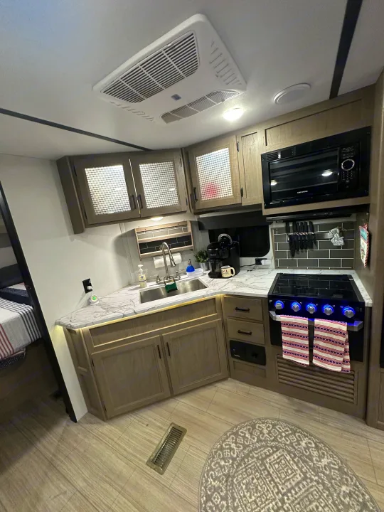 This compact RV rental kitchen features a microwave, sink, stovetop, and light wood cabinets with mesh inserts. Ready for your travels!. Keystone Hideout 2021
