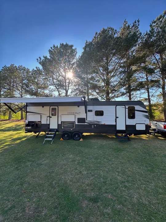 A white & black Hideout travel trailer (RV rental) parked on green grass with awning out, outdoor kitchen visible, under a sunny sky.. Keystone Hideout 2021