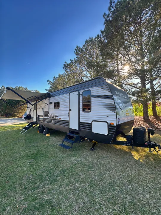 A large travel trailer (RV rental) is parked on green grass under a clear blue sky, with pine trees and sunbeams in the background.. Keystone Hideout 2021