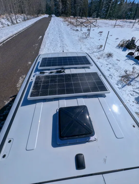 Top view of a white RV rental roof with two solar panels and a vent, parked in a snowy, wooded area on a sunny day.. Dodge Sprinter Van 2015