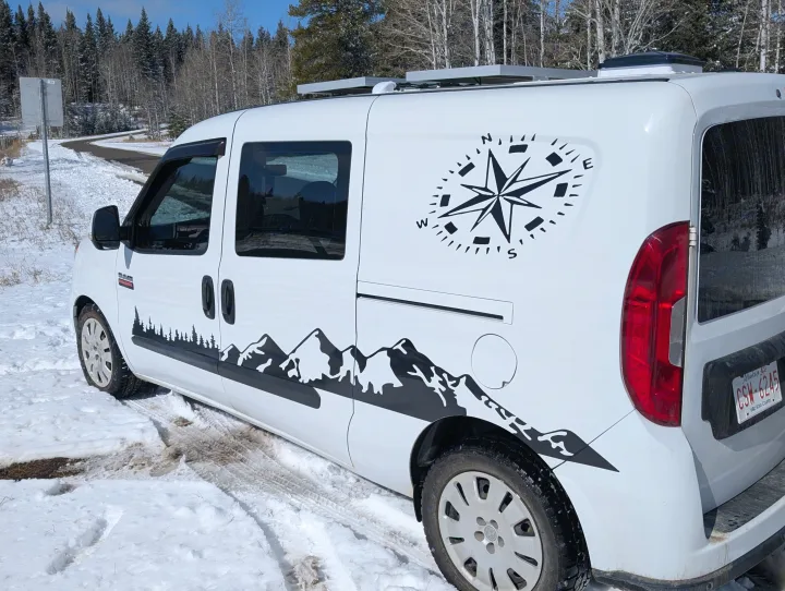 This white custom RV rental, adorned with mountain and compass decals, is parked on a snowy road in a winter landscape.. Dodge Sprinter Van 2015