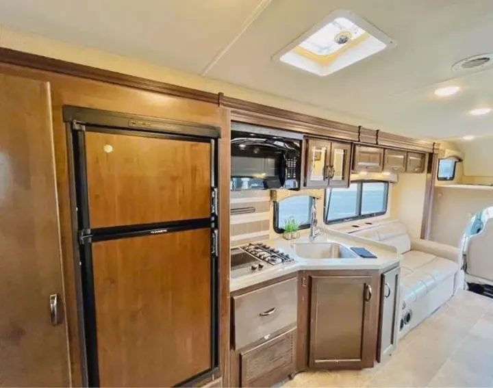 Clean RV rental interior featuring a compact kitchen with stove, sink, microwave, wood-paneled fridge, and comfortable sofa.. Thor Motor Coach Chateau 2019