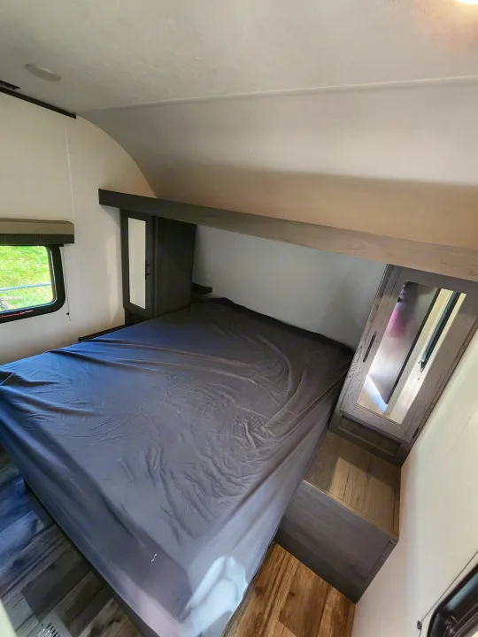 Cozy RV rental bedroom featuring a dark grey bed, overhead storage, and a bright window with green views.. Dutchmen Aspen Trail 26BH 2025