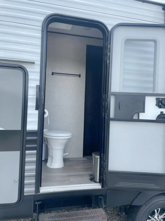 An RV rental's open door reveals a compact bathroom: white toilet, towel bar, and trash can inside the travel trailer.. Jayco Jay Flight 2022