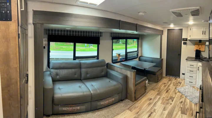 RV rental living space with a comfy gray sofa, dinette, kitchen, and wood-look floor. Large windows reveal outdoor scenery.. Venture Stratus 2021