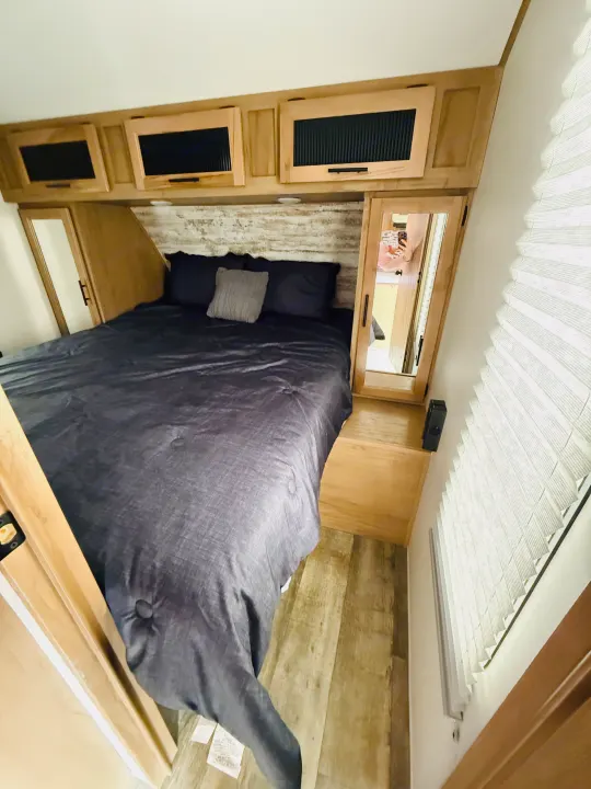 Cozy RV rental bedroom featuring a bed with dark gray bedding, overhead storage, and wood-look flooring and headboard.. Forest River Cherokee Alpha Wolf 2023