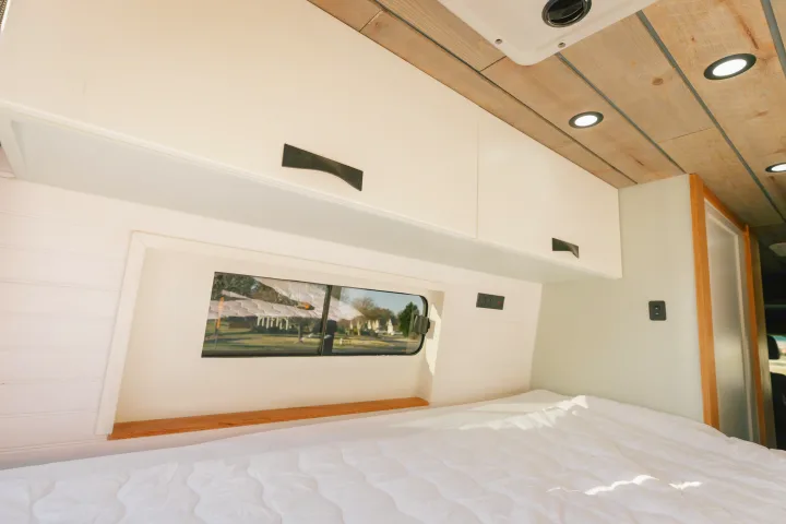 Bright RV rental bedroom features a cozy bed, overhead cabinets, and a window for scenic views.. Sovereign Vans Mercedes Benz Sprinter 2500 4*4 2024