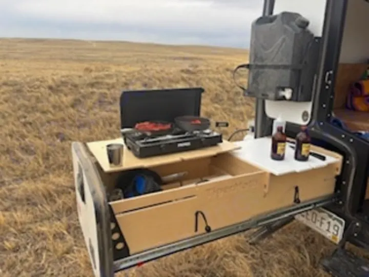 RV rental outdoor kitchen setup extended from vehicle. Cooking and drinks in a grassy field for camping trips.. TAXA Tiger Moth 2023