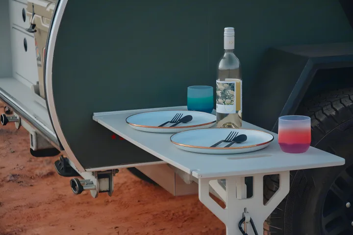 An RV rental's clever pull-out table is set for two with plates, cutlery, a wine bottle, and colorful glasses for outdoor meals.. Escapod Escapod Trailer 2022
