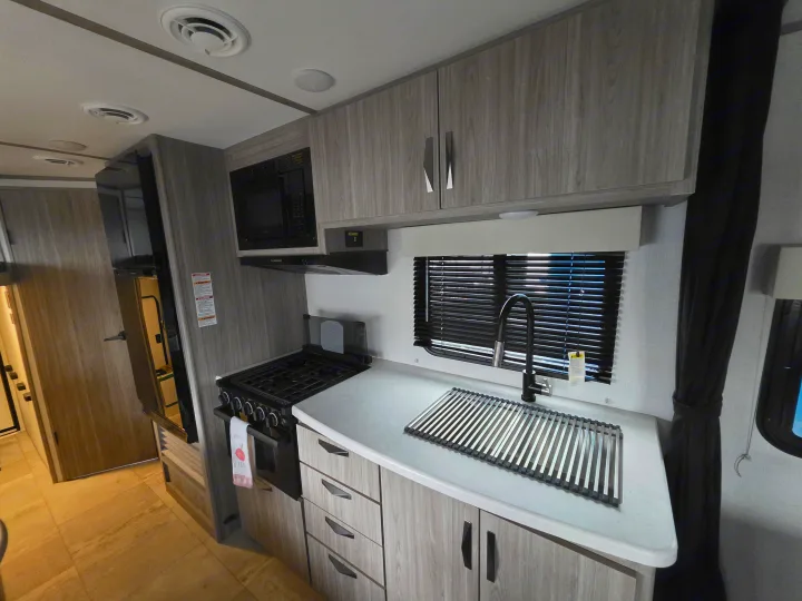 Modern RV rental kitchen featuring light wood-grain cabinets, microwave, stovetop, and sink with a drying rack.. Keystone RV Passport SL Ultra Lite 2024