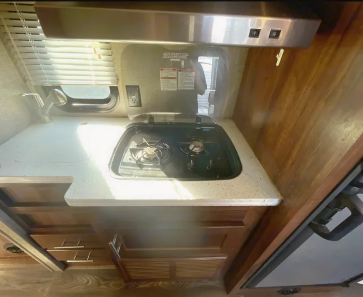RV rental kitchen featuring a two-burner stove, sink, overhead vent, and wood cabinetry. Sun shines through the window.. Heartland Terry Classic V21 2018