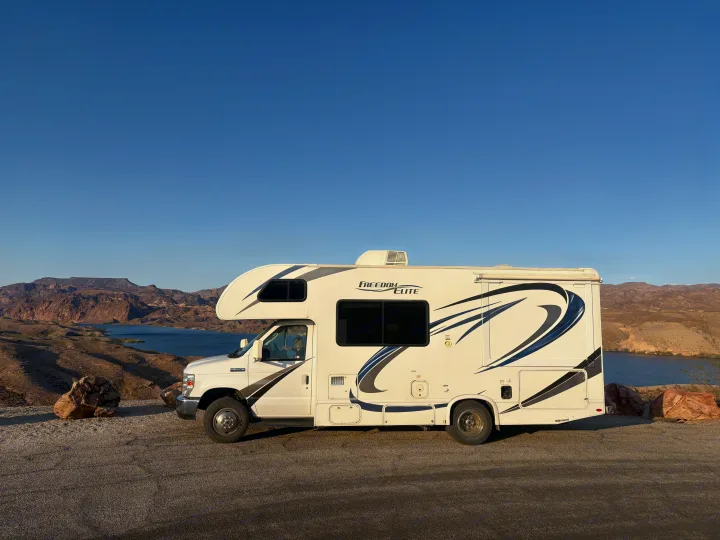 A white Freedom Elite RV rental overlooks a vast lake and rugged mountains under a clear blue sky.. Thor Motor Coach Freedom Elite 2018