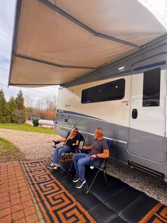 A couple sits in chairs under an extended awning next to their RV rental, on an outdoor mat at a campsite.. Winnebago View 2022