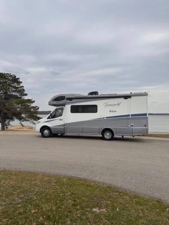 A white and gray Winnebago View Class C motorhome RV rental is parked by a lake on an asphalt road under an overcast sky.. Winnebago View 2022
