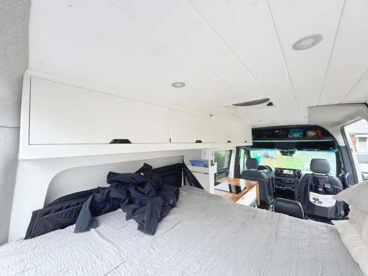 View from the back of an RV rental, showing a grey bed, white overhead cabinets, and a compact kitchen counter, leading to the front cab.. Mercedes-Benz Sprinter 2020