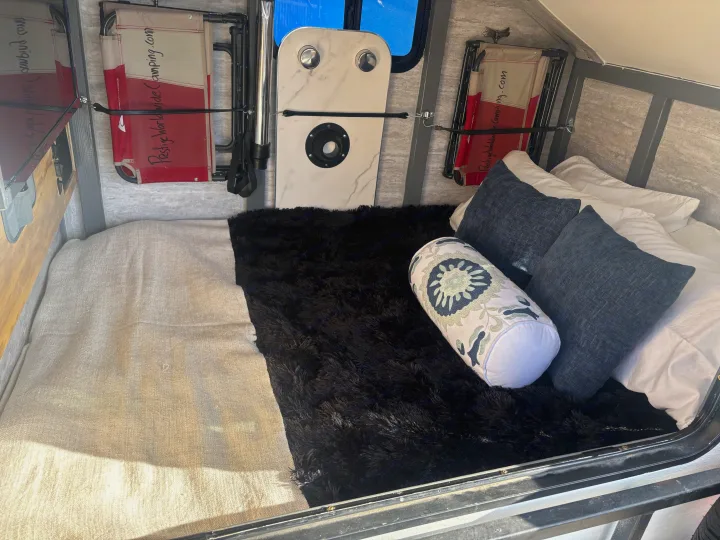 An RV rental interior featuring a bed with pillows, a black fluffy blanket, and wall-mounted folding chairs and a white table.. Overlander HMDE 2025