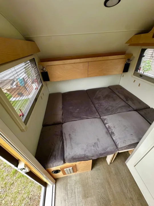 Inside a comfortable RV rental: A plush grey cushioned bed/seating area, overhead wood storage, and bright windows.. nuCamp T@B 2022