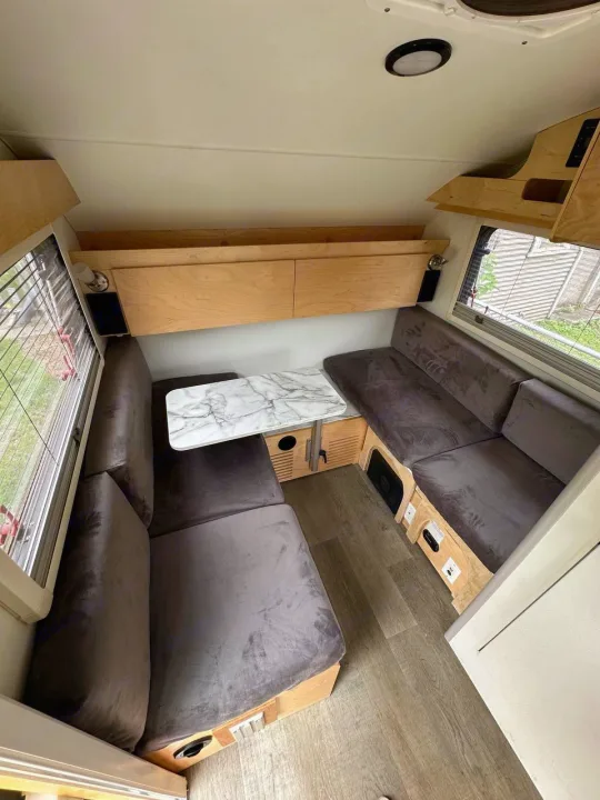 Modern compact RV rental interior showcasing a cozy dinette with gray velvet benches, marble table, and light wood accents.. nuCamp T@B 2022