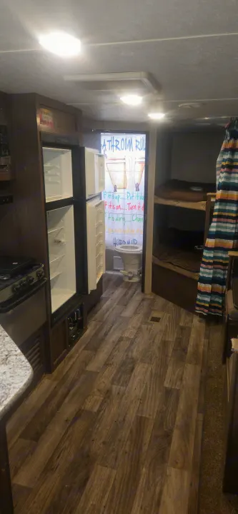 RV rental interior featuring a kitchenette, open fridge, bathroom with "BATHROOM RULES" shower curtain, and bunk beds.. Keystone RV Hideout LHS 2018