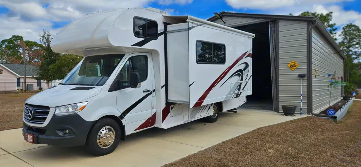 Sleek white Mercedes-Benz RV rental with extended slide-out parked on a driveway beside a large metal garage.. Mercedes Coleman 24CL 2024