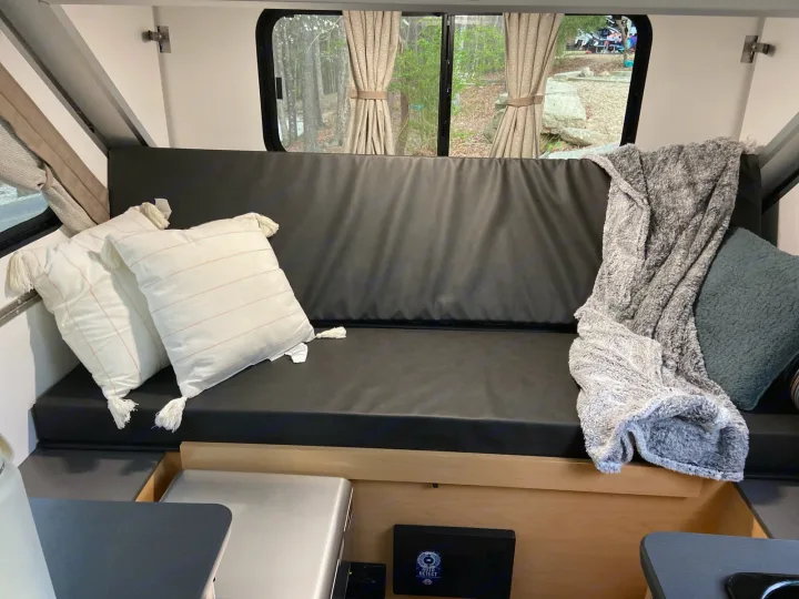 Cozy RV rental interior: black bench/bed, pillows, blanket, and a window view of a serene campground from inside the camper.. A-Liner Ranger 2025
