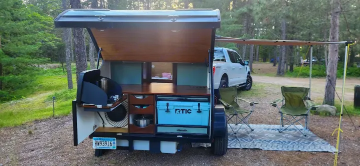 This RV rental teardrop camper features an outdoor kitchen, cozy bed, and shaded setup for serene camping.. Driftwood Custom 2021
