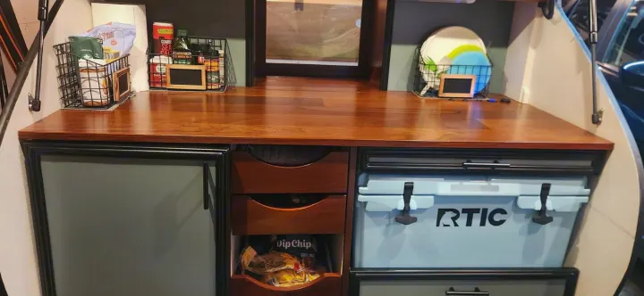 Compact RV rental kitchen with wood counter, RTIC cooler, and organized storage for spices & dishes.. Driftwood Custom 2021