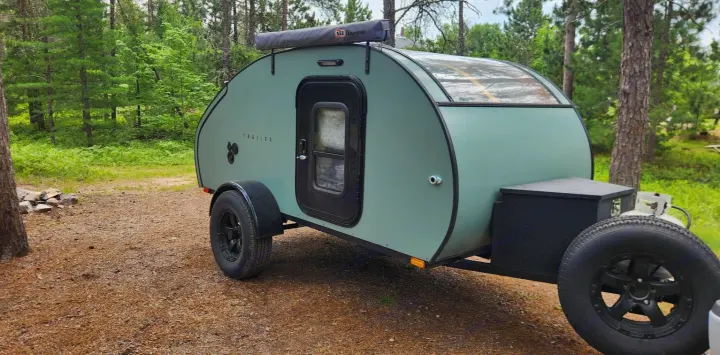 This mint green teardrop trailer, seen in a wooded camp, is ready for your rv rental adventure.. Driftwood Custom 2021