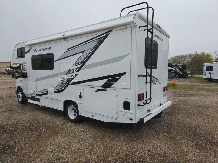 A white Four Winds Class C RV rental with black and grey graphics is parked on a gravel lot, viewed from its rear-side.. Thor Motor Coach Four Winds 2026