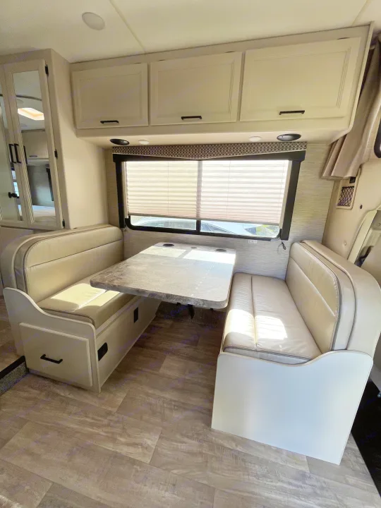 Bright RV rental dinette area with light-colored benches, a gray table, and overhead cabinets, ideal for travel and meals.. Thor Chateau 2023