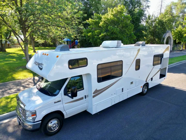 Majestic rv rental: A white Class C motorhome with gold accents, parked next to a green park on a sunny day for travel.. Thor Majestic 2019