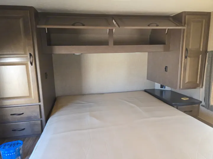 RV rental bedroom: a bed, open overhead storage, left wardrobe with drawers, and right nightstand. Light wood and beige tones.. Thor Majestic 2019