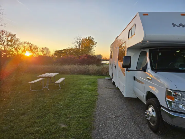 RV rental parked at a scenic campsite with a picnic table, glowing in the light of a vivid sunset over distant trees.. Thor Majestic 2019