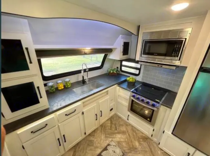 Modern RV rental kitchen interior with white cabinets, dark counters, stainless appliances, sink, and scenic window view.. Forest River Palomino Puma 2021
