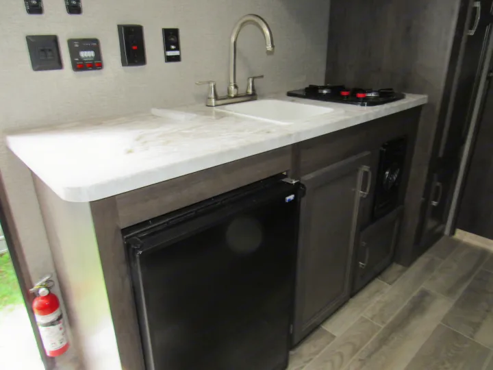 Compact RV rental kitchen with sink, two-burner stove, and mini-fridge. Wall controls are visible above the counter.. Jayco Jay Flight SLX 2021