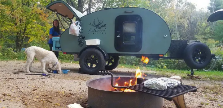 Perfect RV rental getaway: woman and dog enjoy a campfire meal beside a cool teardrop trailer in the woods.. Driftwood Custom 2021