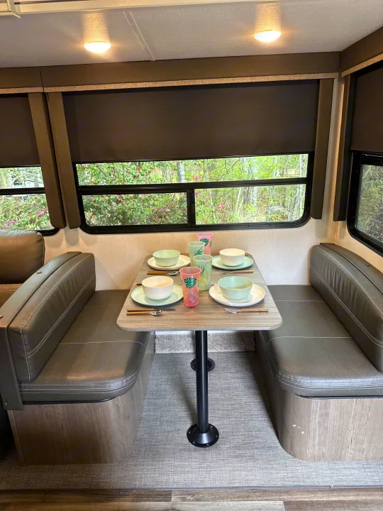 Cozy RV rental dinette with padded benches, a set table for four, and large windows showing trees.. Keystone RV Cougar 2021