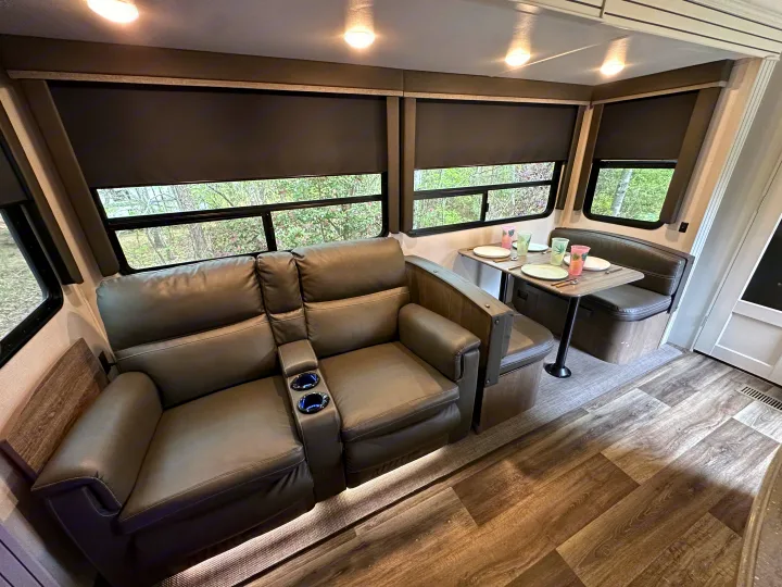 Interior of an RV rental with cozy gray theater seating, a dinette, and large windows showcasing outdoor greenery.. Keystone RV Cougar 2021