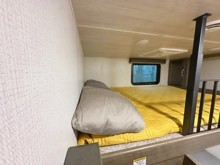 Cozy RV rental bunk bed with a yellow comforter, grey pillow, and window view. Includes AC vent, offering a compact sleeping space.. Keystone RV Cougar 2021