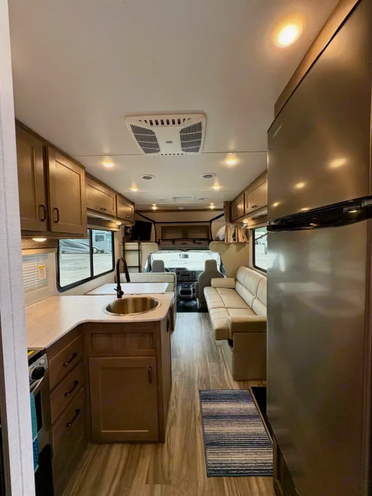 This RV rental interior shows a kitchen with wood cabinets, sink, and stove, a large fridge, and a comfortable sofa/dinette space.. Gulf Stream Conquest 2025