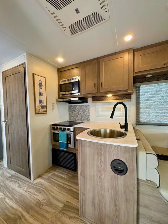 Modern RV rental kitchen with sink, stove, microwave, and dinette, featuring light wood tones and a functional layout.. Gulf Stream Conquest 2025